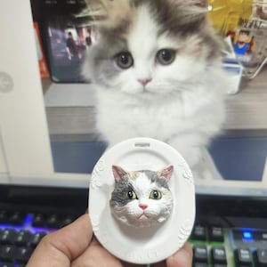 Könnte beinhalten: Ein weißes, ovales Katzen-Ornament mit einem 3D-geformten Katzenkopf. Die Katze hat graues, weißes und braunes Fell, grüne Augen und eine rosa Nase. Das Wort "GUCCI" ist am unteren Rand des Ornaments aufgedruckt.