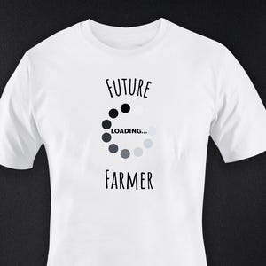 Könnte beinhalten: Weißes T-Shirt mit dem schwarzen Schriftzug "FUTURE FARMER" und einem Ladesymbol in der Mitte. Das Ladesymbol ist eine Reihe von schwarzen und grauen Kreisen. Das Shirt besteht aus einem weichen Material.