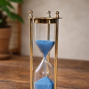 May include: An hourglass with a brass frame and clear glass chambers. The sand inside is a vibrant blue. The hourglass is a decorative item, likely used for timing or as a display piece. The hourglass is approximately 6 inches tall.