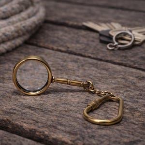 May include: A vintage-style brass magnifying glass keychain with a clear lens, resting on a weathered wooden surface. A small chain connects the magnifier to a carabiner clip. A coil of rope and a set of keys are in the background.