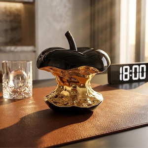 May include: A decorative apple sculpture with a black top and a gold-colored base. The sculpture has a glossy finish and sits on a brown surface. A glass with a sphere inside and a digital clock displaying 18:00 are also visible.