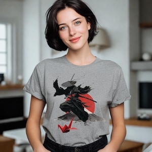 May include: A gray t-shirt featuring a graphic of a samurai warrior in black attire with a red sun and mountain backdrop. A red origami crane is in the foreground. The t-shirt is being worn.