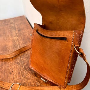 May include: A tan leather crossbody bag with a fold-over flap, visible stitching along the edges, and a zippered pocket on the front exterior. The bag includes an adjustable leather shoulder strap with a silver-toned metal buckle for length adjustment.