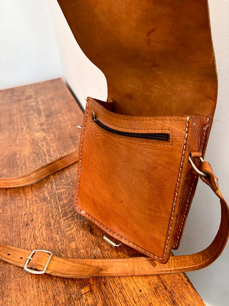 May include: A tan leather crossbody bag with a fold-over flap, visible stitching along the edges, and a zippered pocket on the front exterior. The bag includes an adjustable leather shoulder strap with a silver-toned metal buckle for length adjustment.