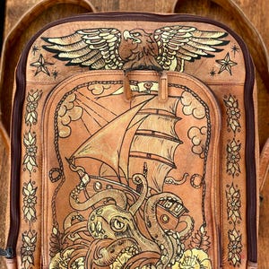 May include: A tan leather backpack featuring intricate embossed and painted designs. The top displays an eagle with spread wings, while the main pocket depicts a sailing ship battling a large octopus, surrounded by floral motifs and stars.