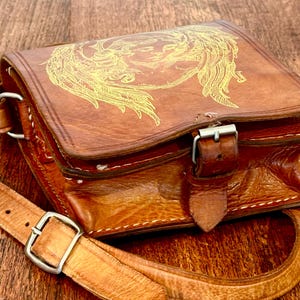 May include: A brown leather satchel featuring a gold-toned line art illustration of a winged figure on the front flap. The bag includes a buckled strap closure, visible white stitching along the edges, and an adjustable shoulder strap with a metal buckle.