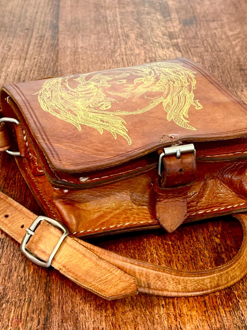May include: A brown leather satchel featuring a gold-toned line art illustration of a winged figure on the front flap. The bag includes a buckled strap closure, visible white stitching along the edges, and an adjustable shoulder strap with a metal buckle.