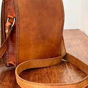 May include: A vertical rectangular brown leather bag featuring a smooth front flap, visible stitched edges, and a long adjustable shoulder strap. The bag is constructed with a textured leather side panel and includes a silver-toned metal hardware ring.