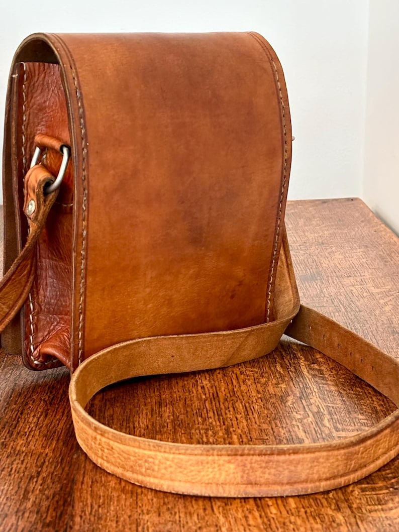 May include: A vertical rectangular brown leather bag featuring a smooth front flap, visible stitched edges, and a long adjustable shoulder strap. The bag is constructed with a textured leather side panel and includes a silver-toned metal hardware ring.
