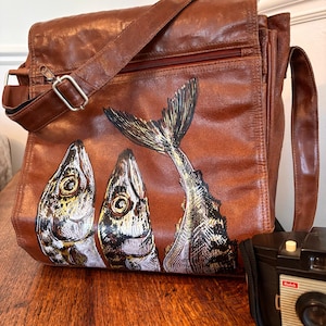 May include: A brown leather messenger bag featuring a graphic illustration of three fish on the front flap. The bag has a zipped pocket, an adjustable shoulder strap with a silver metal buckle, and a black vintage Kodak Brownie Cresta camera.