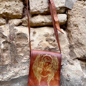 May include: A brown leather crossbody bag featuring a gold-toned engraved illustration of an angel with large wings on the front flap. The bag includes an adjustable leather shoulder strap with a metal buckle and a single leather tab closure.