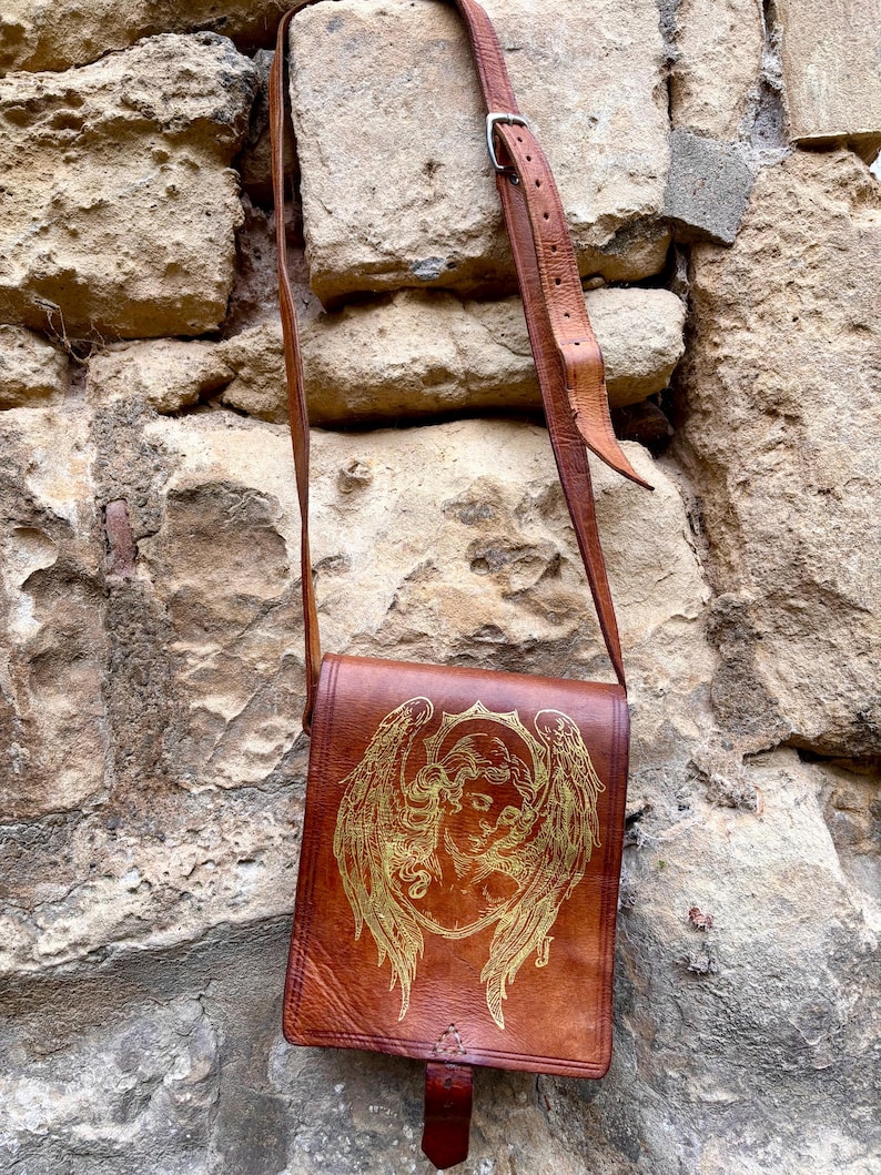 May include: A brown leather crossbody bag featuring a gold-toned engraved illustration of an angel with large wings on the front flap. The bag includes an adjustable leather shoulder strap with a metal buckle and a single leather tab closure.
