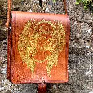 May include: A tan leather shoulder bag featuring a gold-toned line art illustration of an angel with large wings on the front flap. The bag includes a long shoulder strap and a vertical leather closure tab centered at the bottom of the flap.