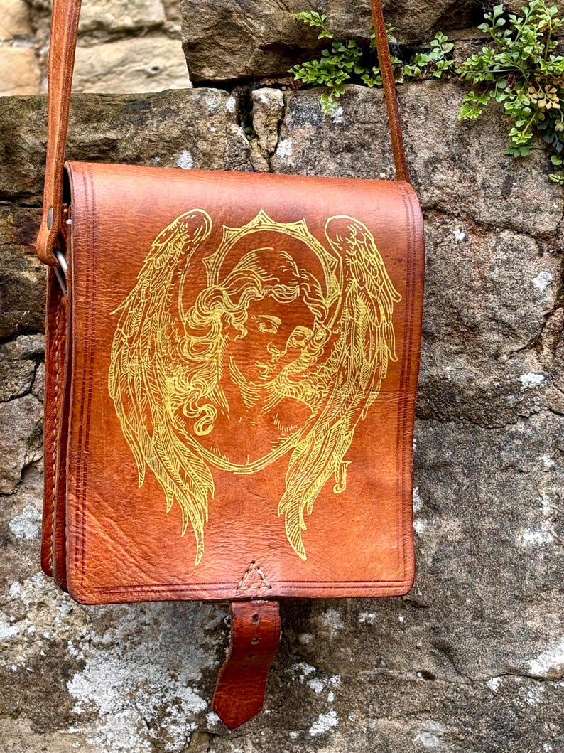May include: A tan leather shoulder bag featuring a gold-toned line art illustration of an angel with large wings on the front flap. The bag includes a long shoulder strap and a vertical leather closure tab centered at the bottom of the flap.