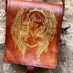 May include: A brown leather satchel featuring a gold-colored line art illustration of an angel with large wings on the front flap. The bag includes a vertical leather strap closure at the bottom center and a long shoulder strap attached to the top.