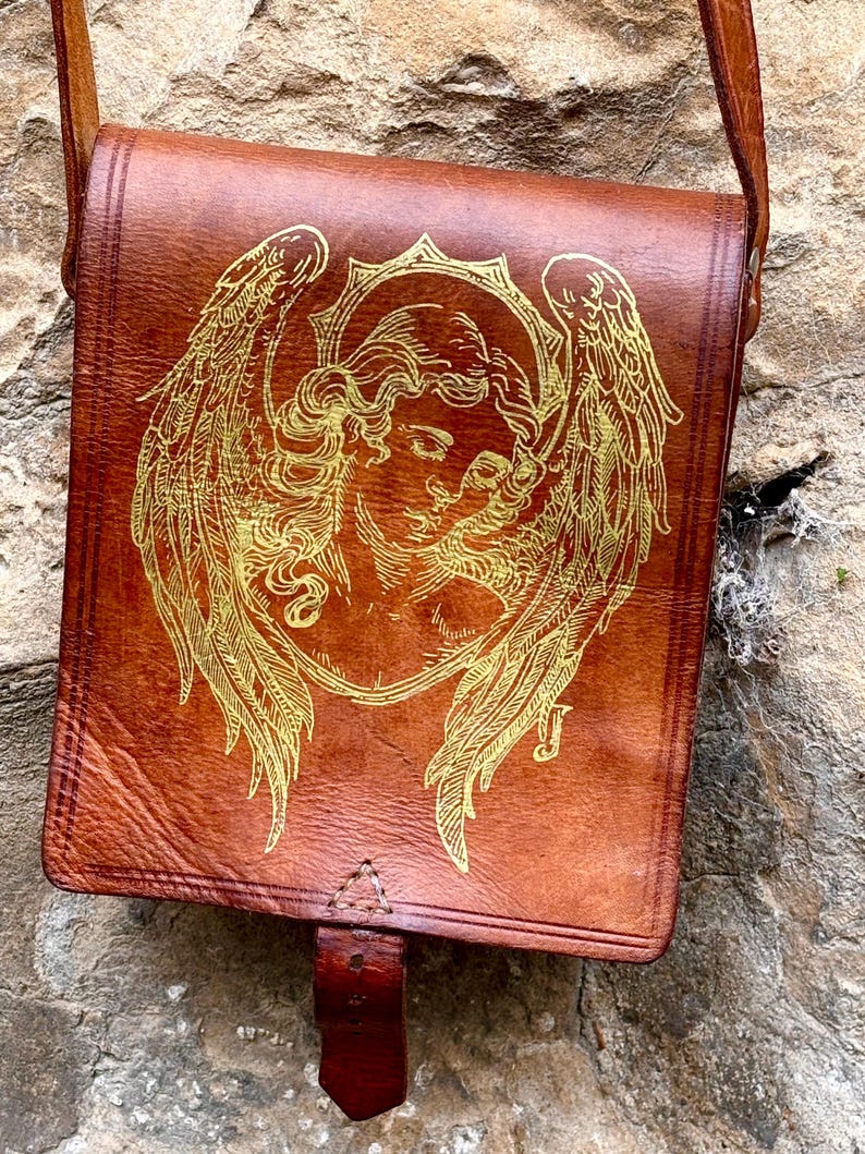 May include: A brown leather satchel featuring a gold-colored line art illustration of an angel with large wings on the front flap. The bag includes a vertical leather strap closure at the bottom center and a long shoulder strap attached to the top.