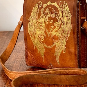 May include: A brown leather shoulder bag featuring a gold-colored line art illustration of an angel with large wings and a halo on the front flap. The bag includes a long, adjustable leather strap with a silver-toned metal buckle for closure.