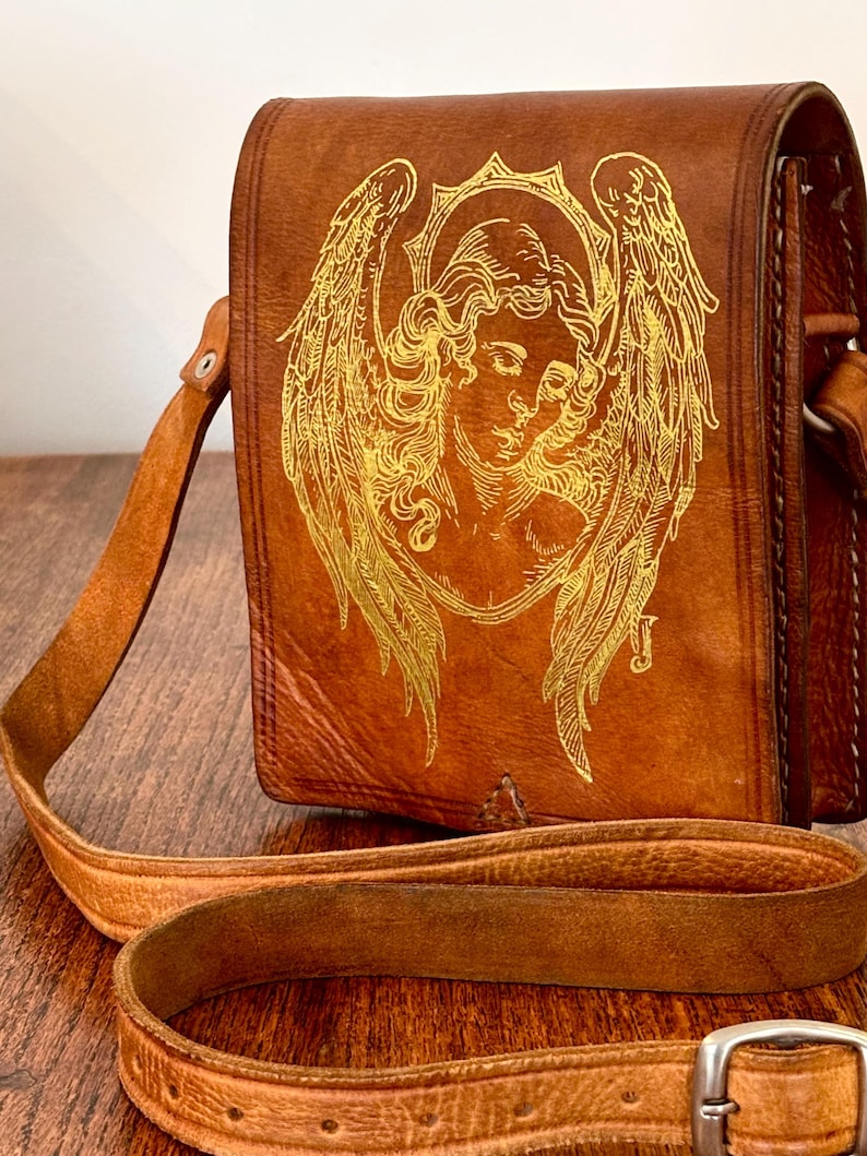 May include: A brown leather shoulder bag featuring a gold-colored line art illustration of an angel with large wings and a halo on the front flap. The bag includes a long, adjustable leather strap with a silver-toned metal buckle for closure.