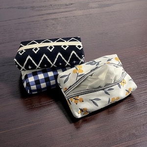 May include: Three fabric tissue box covers in a stack. The top cover is navy blue with a white geometric pattern. The middle cover is blue and white checkered. The bottom cover is white with yellow floral and gray leaf patterns.