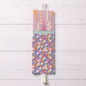 May include: A rectangular fabric bookmark with a pink background and colorful patterns. The top section has a pink background with orange and white decorative lines. The middle section is light blue. The bottom section has a checkered pattern in various colors. A white elastic loop is attached.