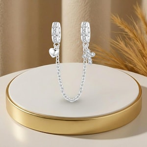 Può includere: Catena di sicurezza per bracciale con ciondoli in argento. Ogni estremità ha un anello ornato di cristalli trasparenti. Un'estremità ha un ciondolo a forma di cuore, l'altra una croce, collegati da una catena. I ciondoli sono su una superficie bianca e dorata.