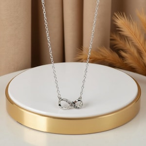 May include: A silver necklace with an infinity symbol pendant, adorned with small, sparkling stones. The necklace is presented on a white and gold platform. The background is neutral in tone.