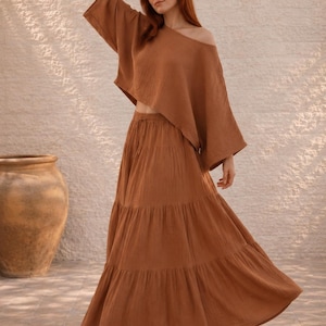 May include: A two-piece brown outfit featuring a cropped, off-the-shoulder top with wide sleeves and a tiered maxi skirt. The outfit is made of a textured fabric and is worn with white flats. A large, brown ceramic pot is in the background.