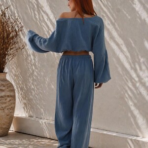 May include: A two-piece blue linen outfit. The top is an off-the-shoulder, long-sleeved crop top with bell sleeves. The pants are wide-legged and high-waisted. The outfit is made of a lightweight, textured fabric. The model is standing in front of a white wall.
