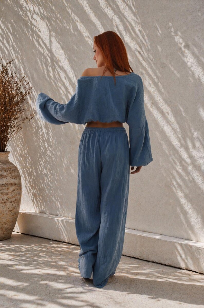 May include: A two-piece blue linen outfit. The top is an off-the-shoulder, long-sleeved crop top with bell sleeves. The pants are wide-legged and high-waisted. The outfit is made of a lightweight, textured fabric. The model is standing in front of a white wall.
