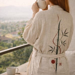 May include: Beige linen robe and shorts set with a bamboo design on the back. The robe has a matching belt and is worn by a person holding a mug. A croissant and fruit are on a nearby table.