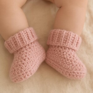 May include: A pair of soft, pink crochet baby booties. The booties have a folded-over cuff and a textured stitch pattern. The image shows the booties on a light-colored surface, suggesting a focus on baby clothing and accessories.