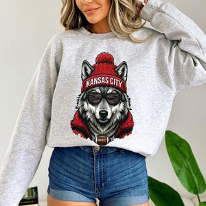 Pu&ograve; includere: Felpa grigio chiaro con un lupo che indossa un berretto rosso con la scritta "KANSAS CITY", occhiali da sole e una giacca rossa. Il lupo ha una catena d'oro con un ciondolo "KANSAS CITY".