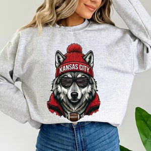 Pu&ograve; includere: Felpa grigio chiaro con un lupo che indossa un berretto rosso con la scritta "Kansas City", occhiali da sole e una giacca rossa. Il lupo ha una catena d'oro con un ciondolo a forma di pallone da football. Girocollo.