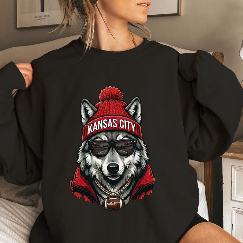 Pu&ograve; includere: Felpa nera con un'illustrazione di un lupo con un berretto rosso con la scritta "Kansas City", occhiali da sole e una giacca rossa. Il lupo ha una catena d'oro con un ciondolo a forma di pallone da football con "Kansas City".