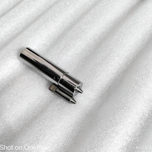 Peut inclure: Gros plan sur deux pièces de machine métalliques et brillantes. La plus grande est cylindrique avec une extrémité pointue et une petite pièce rectangulaire attachée. Les pièces sont sur une surface blanche texturée.