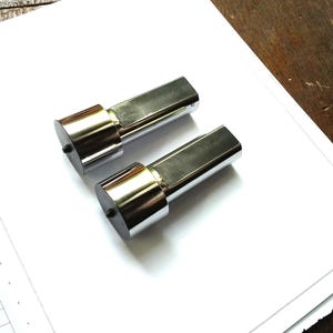 Könnte beinhalten: Zwei glänzende, metallisch-silberne Objekte mit zylindrischem Sockel und rechteckiger Oberseite. Die Objekte liegen auf einer weißen Oberfläche, möglicherweise Papier, mit einem Holzgrund.