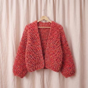 Könnte beinhalten: Ein grob gestrickter Cardigan in leuchtendem Rot, an einem Holzbügel aufgehängt. Das handgestrickte Kleidungsstück hat einen strukturierten Stich und lange Ärmel und bietet eine gemütliche und stilvolle Ästhetik. Der Hintergrund ist eine weiche, neutrale Farbe.