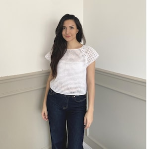 May include: A white, open-weave crochet top with short sleeves. The top has a round neckline and a loose fit. The model is wearing dark blue jeans. The top is a lightweight, breathable garment, suitable for warmer weather.