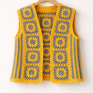 Puede incluir: Un chaleco hecho a mano, sin mangas, con un patrón de cuadrados de la abuela. El chaleco presenta una combinación de cuadrados amarillos y grises, con rayas amarillas en los laterales. El chaleco se muestra en una percha de madera sobre un fondo blanco.