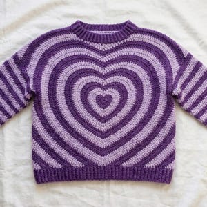 May include: A purple and lavender crochet sweater featuring a large heart design on the front. The heart is made of concentric stripes, with a ribbed collar and cuffs. The sleeves are long and striped.
