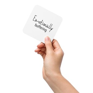 Puede incluir: Una mano sostiene una tarjeta cuadrada blanca con la inscripción manuscrita en negro «Emotionally buffering». La tarjeta está ligeramente inclinada, sobre un fondo blanco.