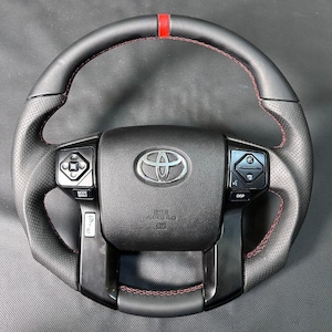 Pode incluir: Um volante de couro preto para veículo Toyota com pegas laterais perfuradas, costuras vermelhas em contraste e uma marca central vermelha no topo. O cubo central exibe o logótipo da Toyota e inclui botões de controlo integrados.