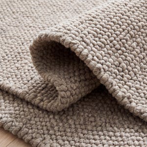 May include: A beige area rug featuring a thick, chunky, hand-knotted texture with a dense, raised loop pile construction. The rug is folded over to reveal the consistent, uniform, and rounded knot pattern across the entire surface area.