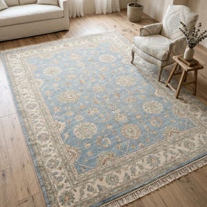 Puede incluir: Una alfombra rectangular que presenta un campo central azul claro adornado con intrincados motivos florales y de enredaderas en tonos crema, beige y salvia suave. La alfombra está enmarcada por un borde ancho color crema con patrones florales a juego y bordes con flecos.