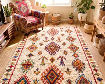 May include: A cream-colored, rectangular area rug with a vibrant geometric pattern in red, blue, yellow, and orange. The rug has a fringed edge and is placed in a room with a patterned armchair and plants.