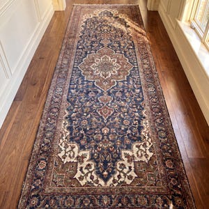 May include: A long, rectangular runner rug featuring a deep blue background with intricate floral and vine patterns in shades of cream, brown, and muted red. The rug has a central medallion design and a detailed border with fringe at both ends.
