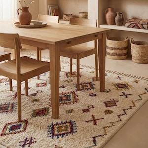 May include: A light wood dining table with four matching chairs. A woven rug with geometric patterns in red, blue, yellow, and green covers the floor. Decorative baskets and pottery are on a shelf in the background.