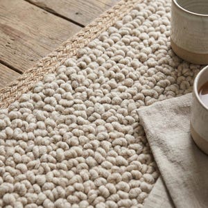 Puede incluir: Un camino de mesa beige texturizado con un borde de yute trenzado. El camino de mesa tiene un tejido grueso y rizado. Dos tazas de cerámica y una servilleta de lino están colocadas sobre él, creando un ambiente de tonos neutros.
