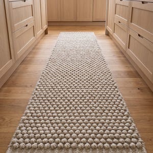 May include: A long, rectangular runner rug featuring a textured, raised bobble pattern in a neutral beige color. The rug is constructed with a dense, woven base and a repeating series of soft, rounded tufts covering the entire surface area.