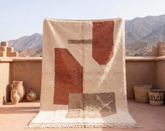 May include: A large, rectangular rug with a cream-colored background and geometric shapes in shades of rust, brown, and gray. The rug has a fringe along the bottom edge. The rug is displayed outdoors, with mountains in the background.